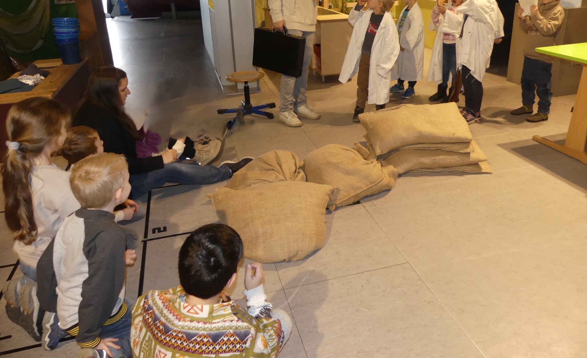 Kinder sitzen im Museum auf dem Boden und schauen auf die Leinensäcke und anderen Gegenstände.
