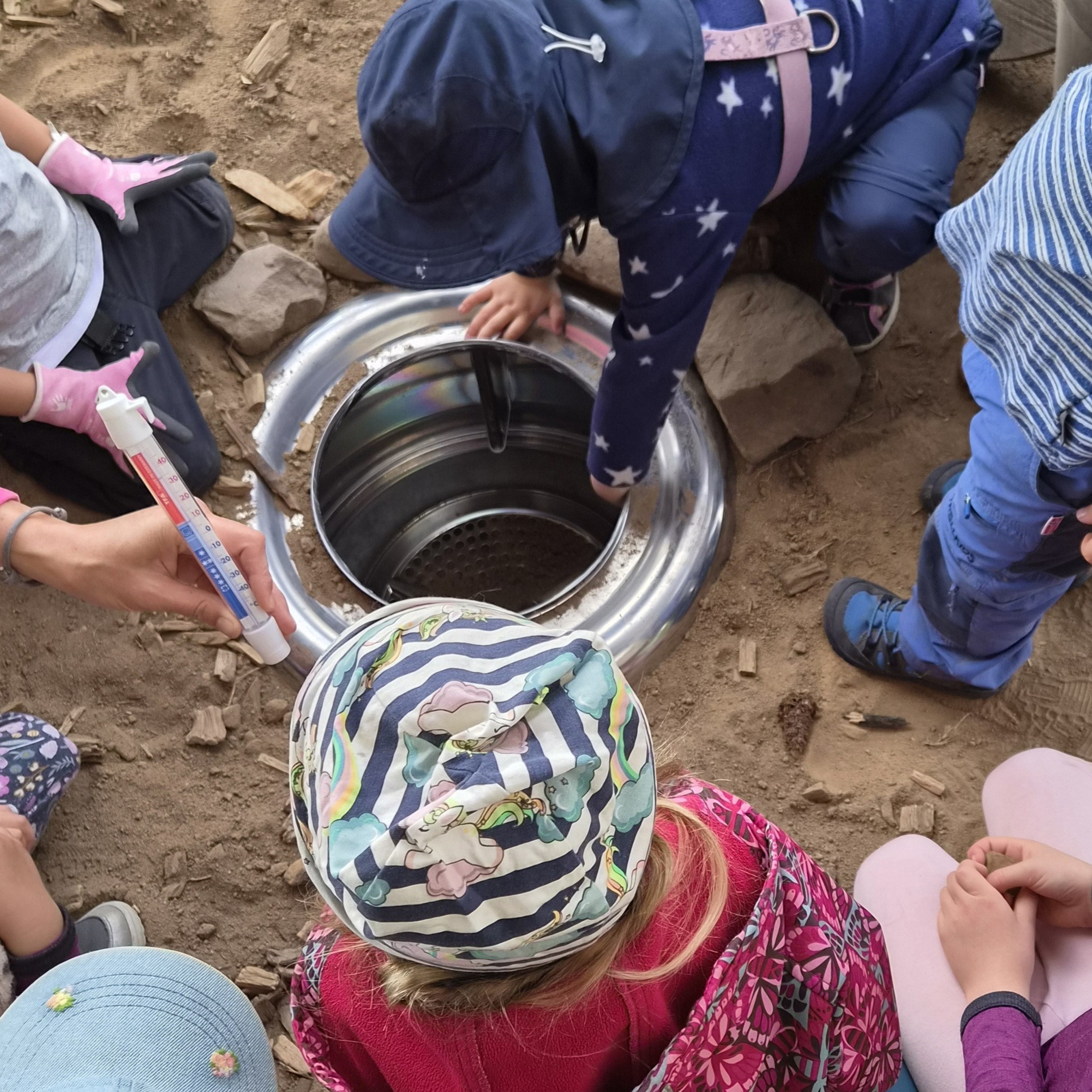 Kinder untersuchen den neuen, eingegrabenen Erdkühlschrank aus einer alten Waschmaschinentrommel.